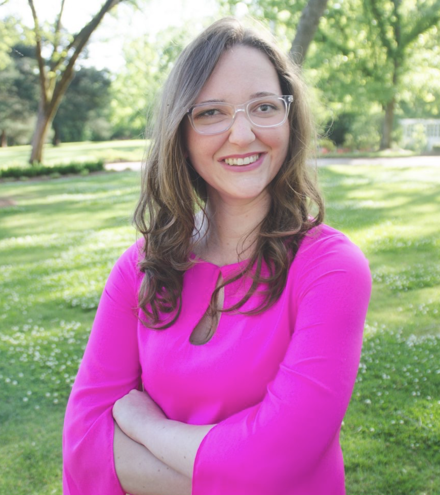 Picture of a young woman wearing glasses, smiling at the camera, wearing a bright pink dress.